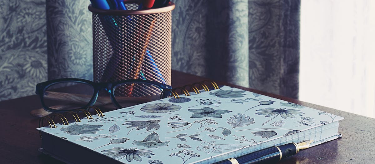 A stylish workspace featuring a floral notebook, glasses, and pens on a wooden desk.