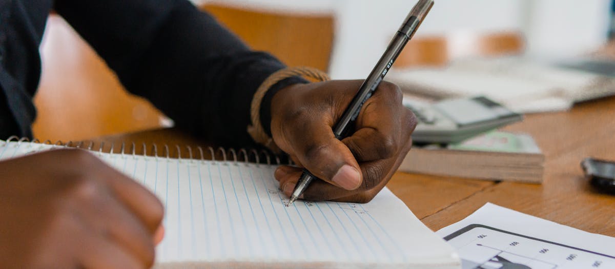 Close-up of hand writing on notebook at a wooden table, featuring paper and ballpen.